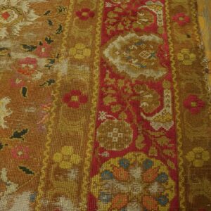 Heavily textured, worn rug in mustard and red floral pattern rests beside yellow hardwood flooring.
