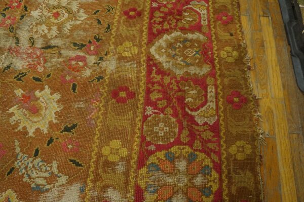 Heavily textured, worn rug in mustard and red floral pattern rests beside yellow hardwood flooring.