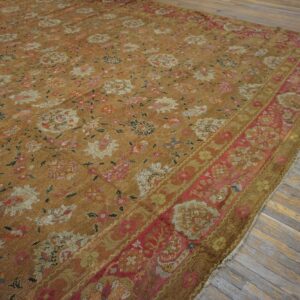 Low-pile rug with a muted gold field, multicolored floral pattern, and a wide pink border on wood floorboards.