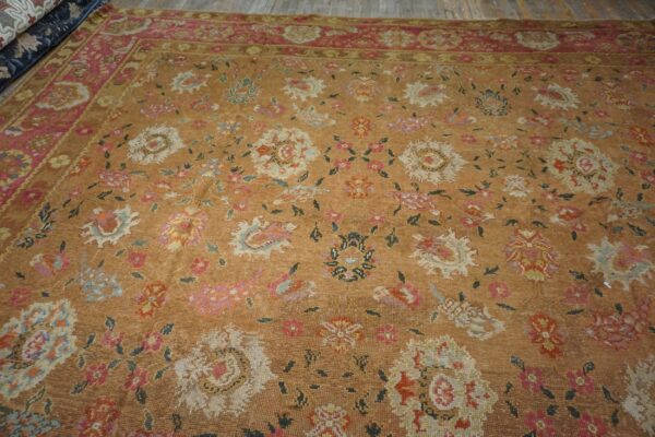 Camel rug with multicolored floral design and wide pink border resting on wood planks.