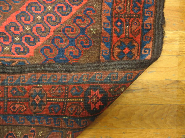 The corner of a red and blue patterned rug, showing the dense pile and woven reverse on wood floors.