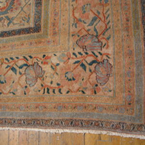 Corner detail of a muted gold rug with intricate blue, red, and tan floral patterning on hardwood floors.