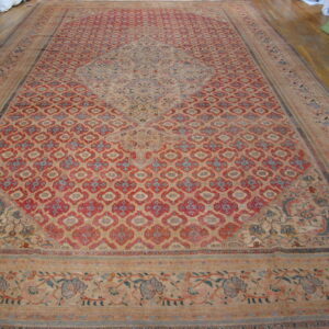 A very large faded rug with a red patterned field, blue medallion, and beige borders on wood floor.