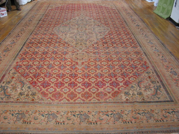 A very large faded rug with a red patterned field, blue medallion, and beige borders on wood floor.