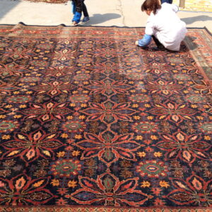A large dark blue rug with repeating red floral motifs lies on concrete outdoors near a person.