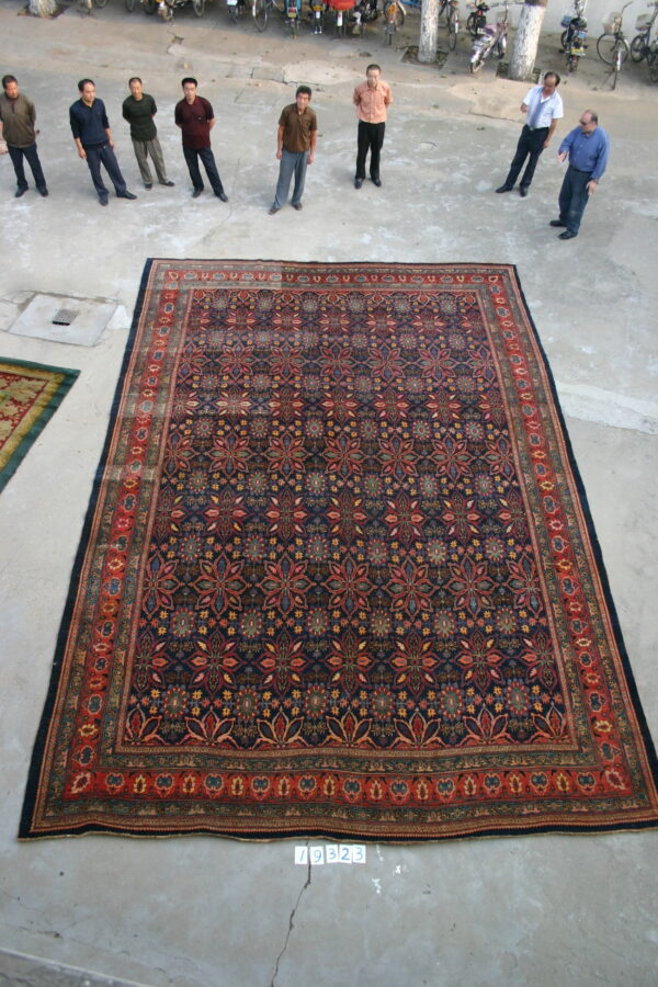A large dark blue patterned rug with complex red borders rests on concrete, viewed by nine people outdoors.