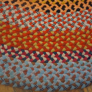 Multicolored braided rug featuring concentric rings of orange, red, yellow, and blue fabric patterns on a wood floor.