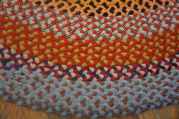 Multicolored braided rug featuring concentric rings of orange, red, yellow, and blue fabric patterns on a wood floor.