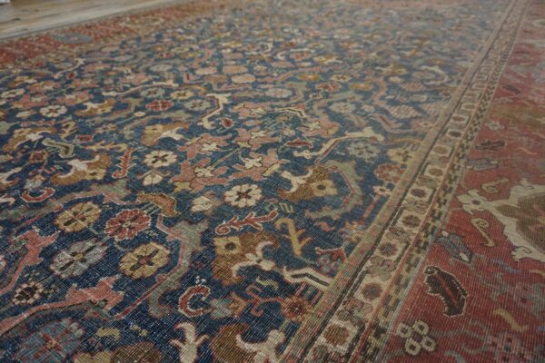Traditional blue rug featuring detailed floral motifs in faded pink, beige, olive, and a wide red border.