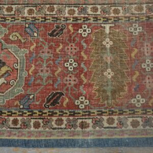 Intricate, low-pile rug with red, green, and cream floral and geometric patterns shown against wood flooring.