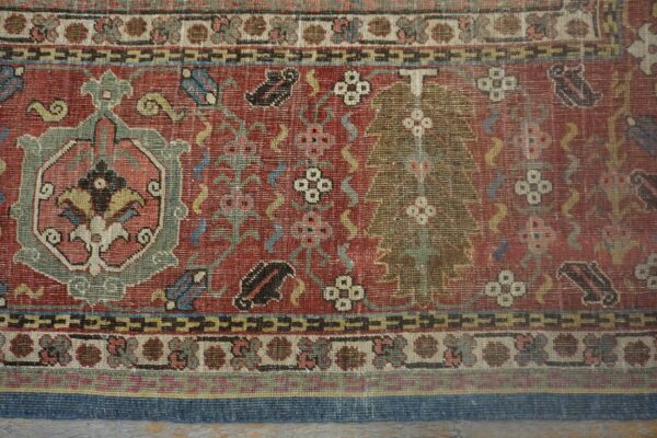 Intricate, low-pile rug with red, green, and cream floral and geometric patterns shown against wood flooring.