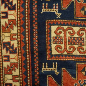 Detail of a low-pile rug featuring a navy blue field and intricate red and cream geometric borders on wood flooring.