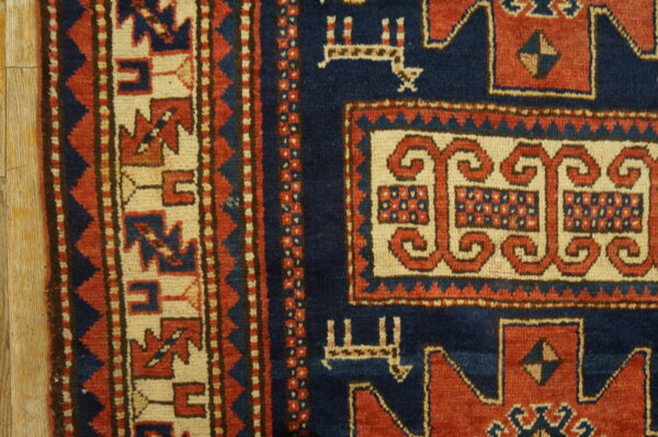 Detail of a low-pile rug featuring a navy blue field and intricate red and cream geometric borders on wood flooring.