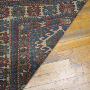A corner of a low-pile rug with geometric patterns in blue, red, and beige lies on light wood flooring.