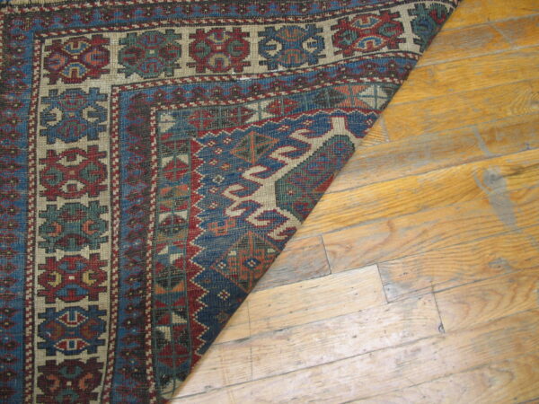 A corner of a low-pile rug with geometric patterns in blue, red, and beige lies on light wood flooring.