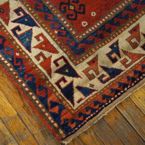 A low-pile rug corner featuring red, navy, and tan geometric patterns on distressed wood flooring.