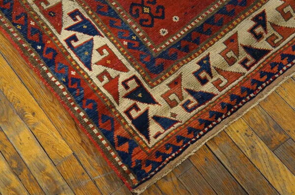 A low-pile rug corner featuring red, navy, and tan geometric patterns on distressed wood flooring.