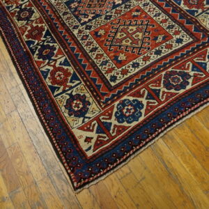 A low pile geometric rug in red, navy, and cream rests diagonally on worn plank hardwood flooring.