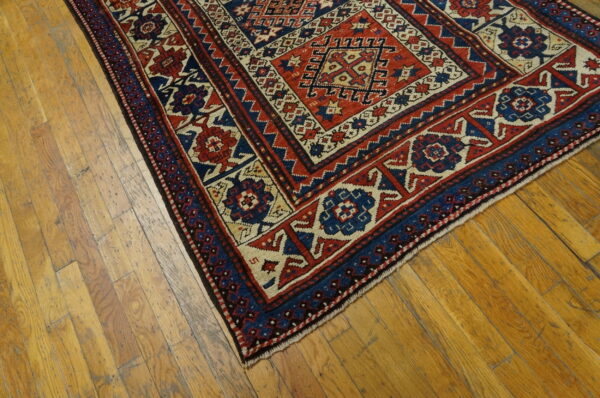 A low pile geometric rug in red, navy, and cream rests diagonally on worn plank hardwood flooring.