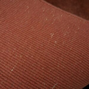 Reddish-brown rug detail showing parallel diagonal ribs and scattered light-colored inclusions.