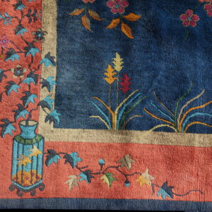 Blue rug with orange and tan borders, featuring stylized floral vines and a decorative vase design.