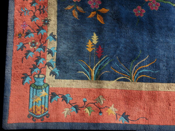 Blue rug with orange and tan borders, featuring stylized floral vines and a decorative vase design.