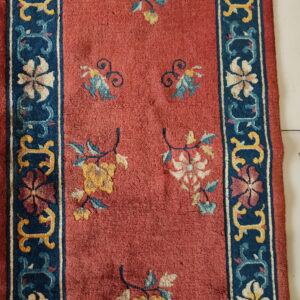 Runner rug with a rust-red field and navy border featuring floral motifs rests on white tiled flooring.