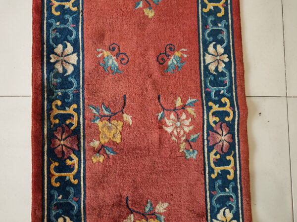 Runner rug with a rust-red field and navy border featuring floral motifs rests on white tiled flooring.