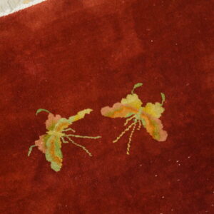 Deep red rug with green and yellow stylized insect patterns next to distressed light wood flooring.