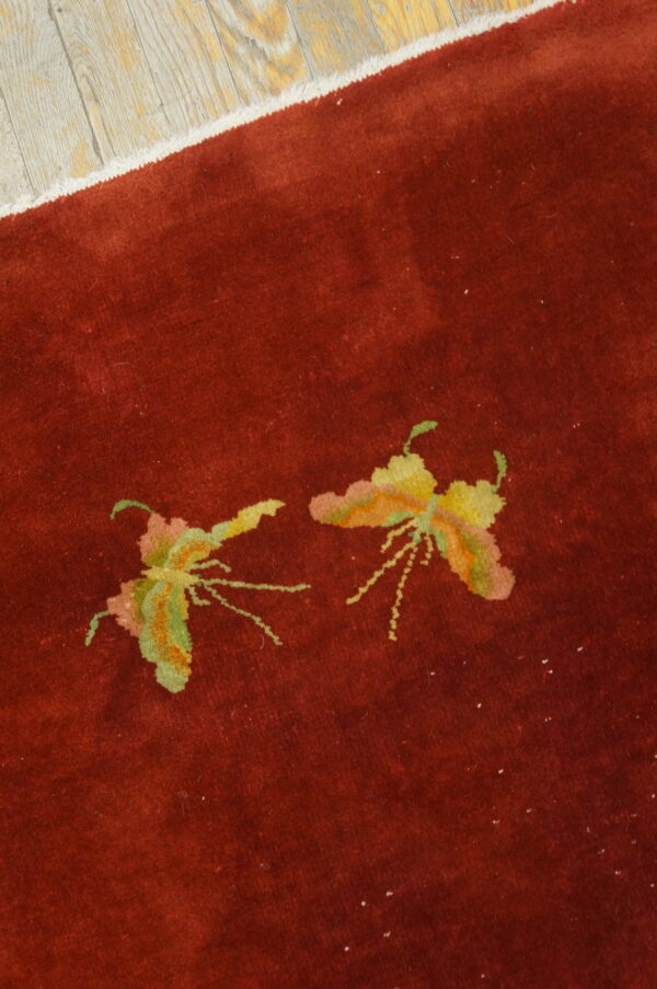 Deep red rug with green and yellow stylized insect patterns next to distressed light wood flooring.