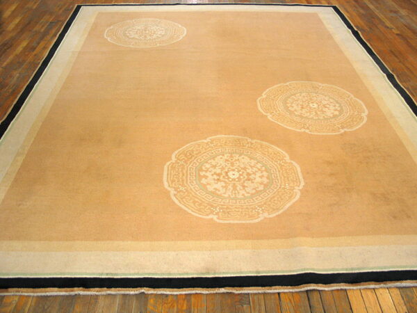 Large tan rug with three pale medallions and a striped border, lying flat on wide plank hardwood flooring.