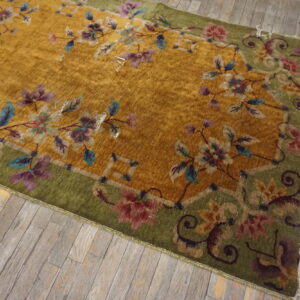Gold low-pile rug with a decorative green border featuring multi-colored floral designs on distressed wood.