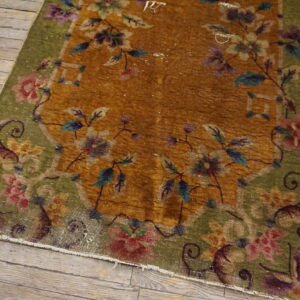 A low-pile floral rug featuring a golden-brown field and a green border rests on rustic plank wood flooring.