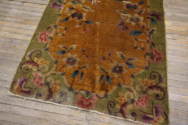 A low-pile floral rug featuring a golden-brown field and a green border rests on rustic plank wood flooring.
