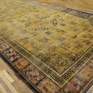 Faded gold rug with scattered black designs and a complex meander and floral border on wood planks.