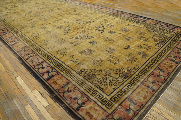 Faded gold rug with scattered black designs and a complex meander and floral border on wood planks.