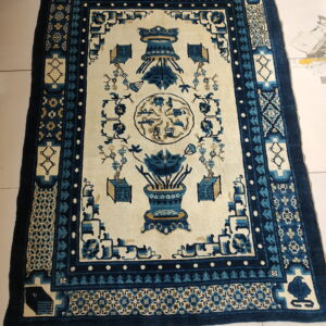 Rectangular rug with an ivory center and elaborate blue and tan stylized botanical patterns on a tile floor.