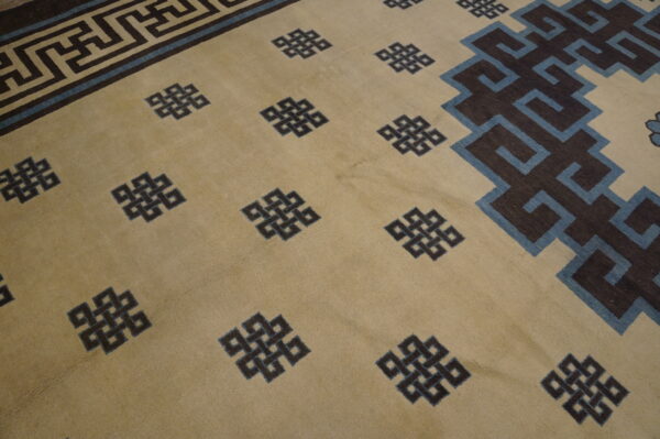 Tan rug with scattered dark knots, blue accents, and a wide geometric dark brown meander border.