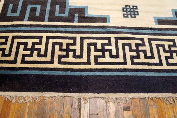 Black, cream, and blue rug border featuring a geometric meander pattern resting on exposed wood planks.