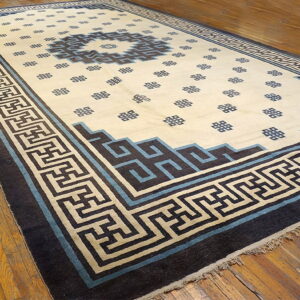 Low-pile rug in cream, blue, and brown, featuring a geometric pattern and wide key border on wood floors.