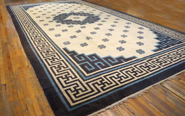 Low-pile rug in cream, blue, and brown, featuring a geometric pattern and wide key border on wood floors.