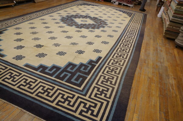 Low-pile rug with geometric patterns in cream, dark brown, and light blue on worn wood flooring.
