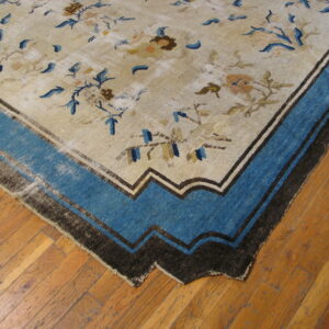 Low-pile beige rug with faded blue floral motifs and a wide blue and dark brown stepped border on hardwood.