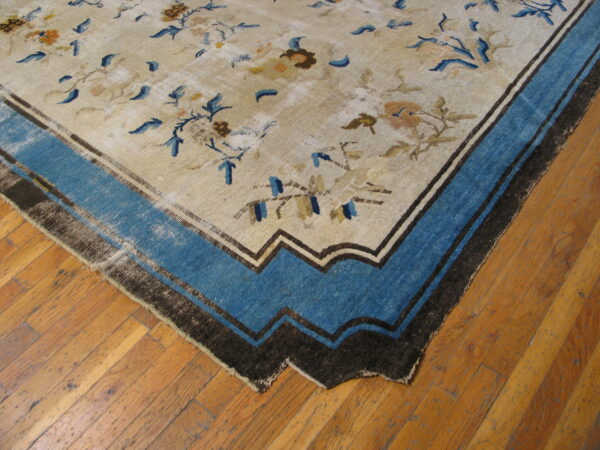 Low-pile beige rug with faded blue floral motifs and a wide blue and dark brown stepped border on hardwood.