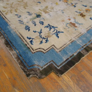 A light-colored floral rug with a prominent blue and dark brown stepped border is laid on wood planks.