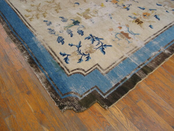 A light-colored floral rug with a prominent blue and dark brown stepped border is laid on wood planks.