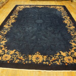 Large navy rug with tone-on-tone field and thick gold and orange floral border on a yellow wood floor.