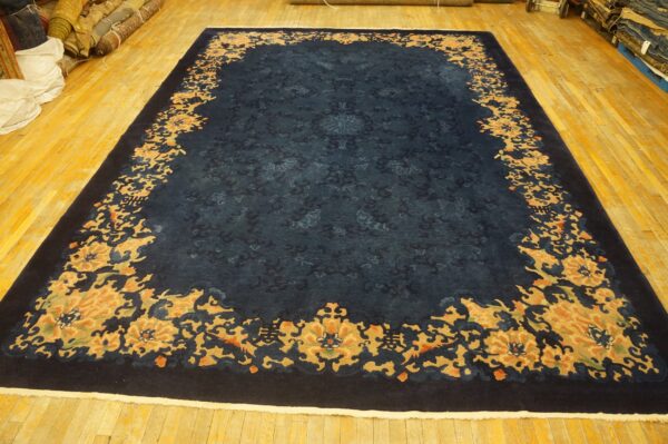 Large navy rug with tone-on-tone field and thick gold and orange floral border on a yellow wood floor.