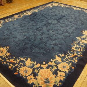Large dark blue rug with repeating stylized tan and blue floral motifs on light hardwood flooring.