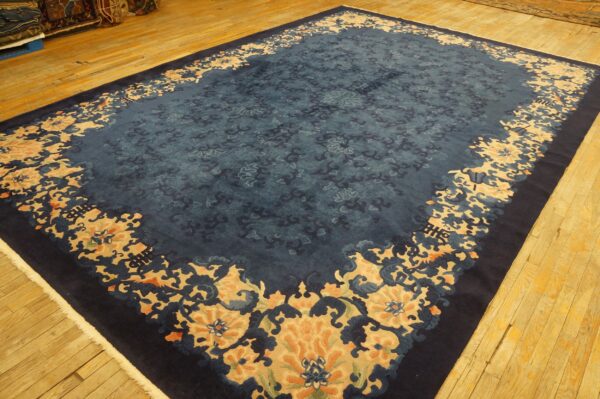 Large dark blue rug with repeating stylized tan and blue floral motifs on light hardwood flooring.